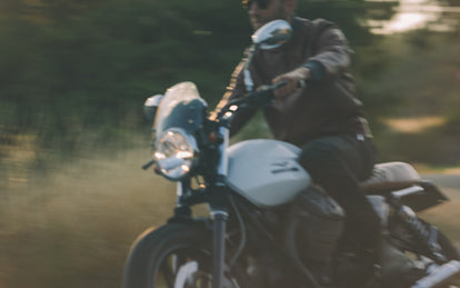 Blurred shot of a man riding a motorcycle, wearing a brown leather jacket.