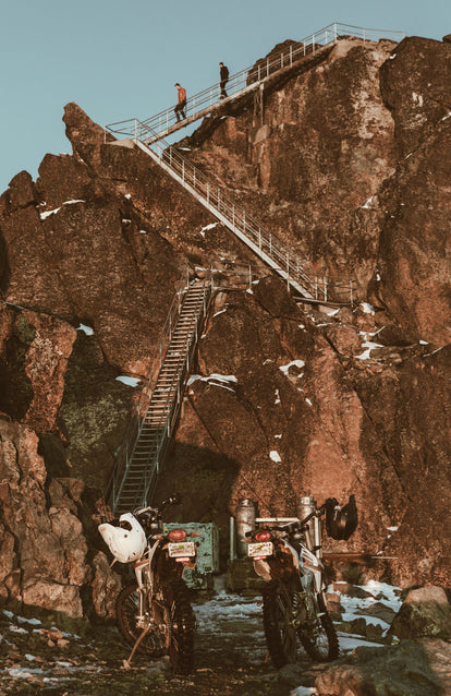 Two guys descending several flights of metal steps down a cliff, towards their waiting motorcycles.