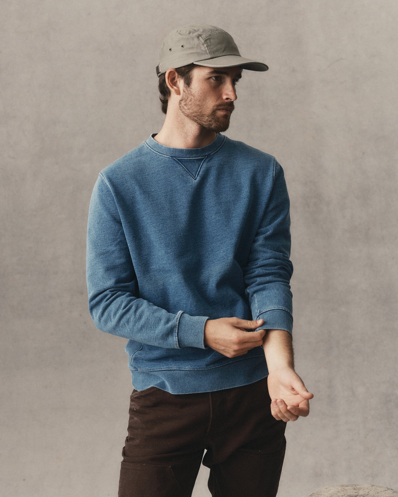A man wearing a heathered blue crewneck sweatshirt, dark brown pants, and a gray baseball cap, standing against a textured gray wall., Knits by Taylor Stitch