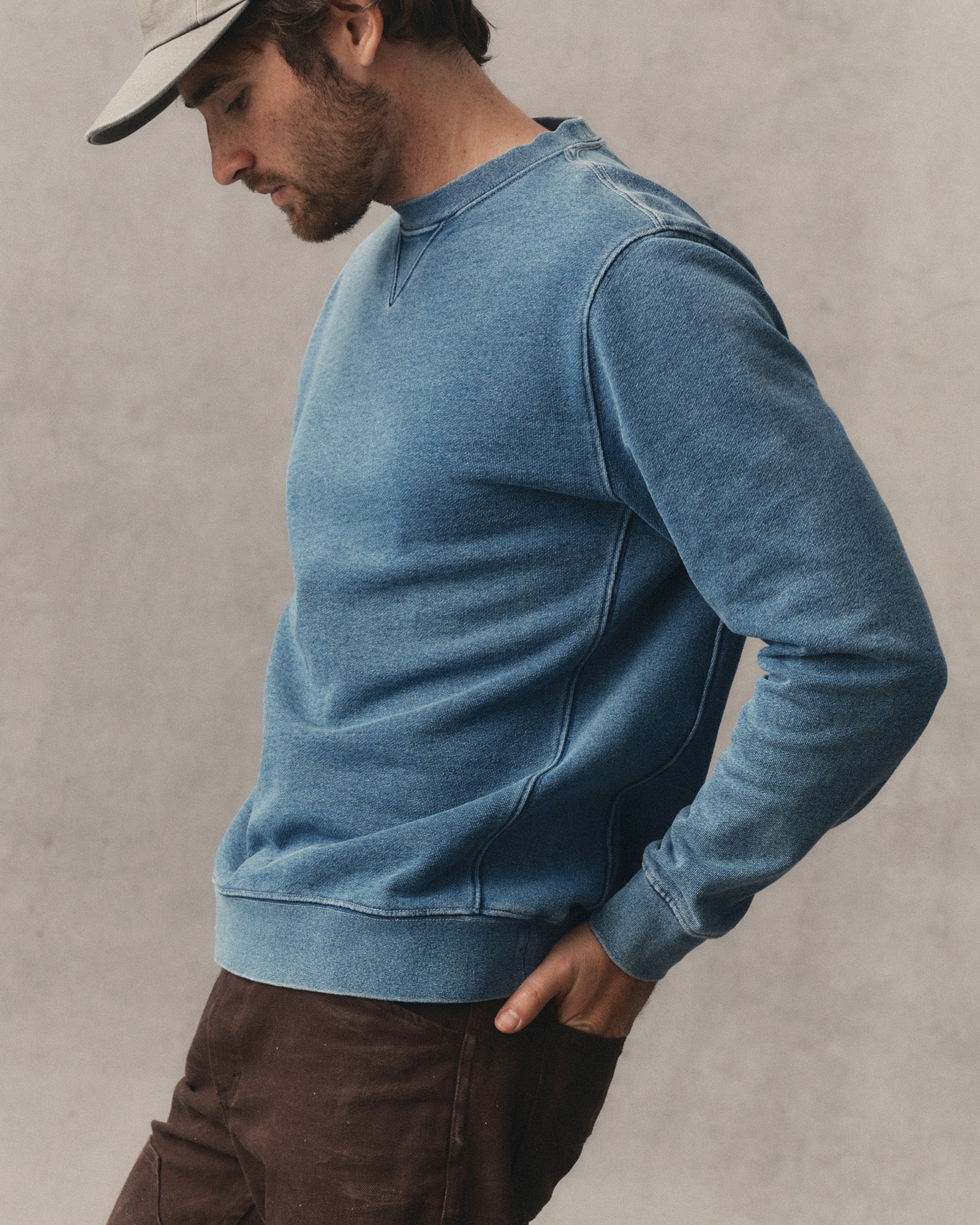 A man wearing a faded medium-blue crewneck sweatshirt and brown pants, shown in a side profile against a textured gray wall., Knits by Taylor Stitch