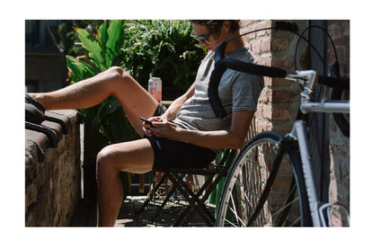Ryan sitting with his feet up on a wall, hydrating and checking his phone with his bicycle leaning against the wall beside him.
