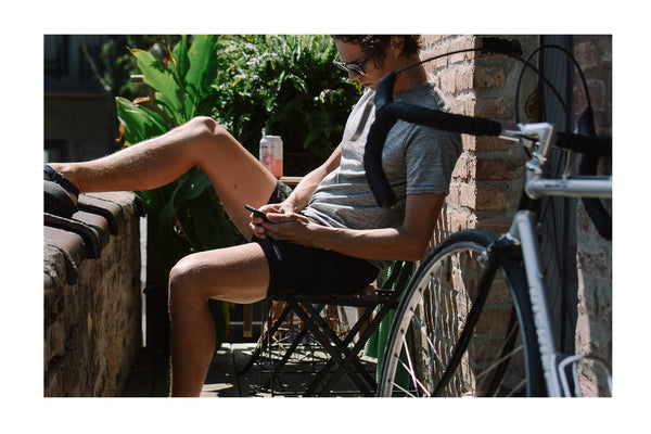 Ryan sitting with his feet up on a wall, hydrating and checking his phone with his bicycle leaning against the wall beside him.