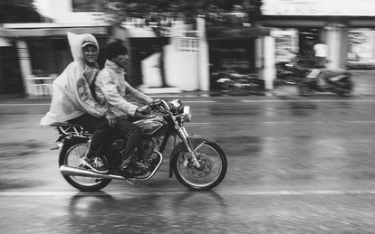 A black and white action shot of two local men travelling down a rainy street on a motorcycle.