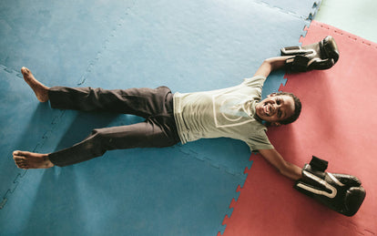 Overhead shot of a young boy lying barefoot on a colored gym mat, wearing large boxing gloves.
