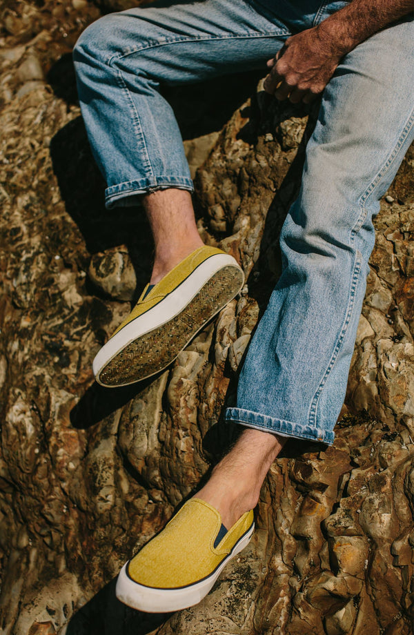 Close-cropped shot of our man sitting on some rocks cross-legged in a pair of yellow pumps, jeans, no socks.
