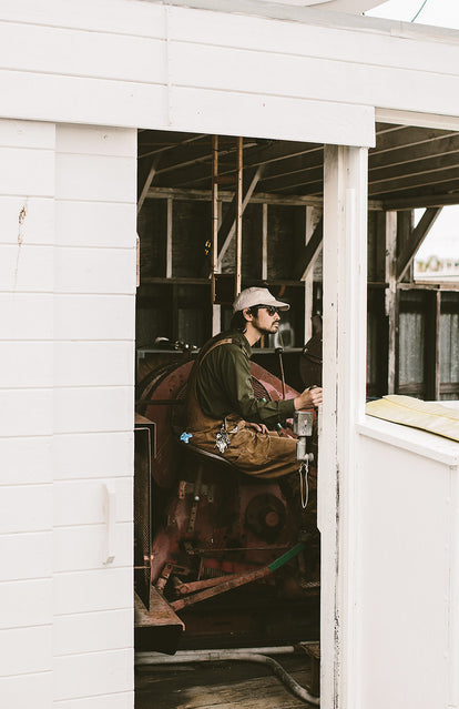 An overalled guy wearing a cap and sunglasses, operating some kind of equipment inside a white-washed wooden building.