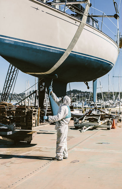 A man wearing PPE, roller painting blue anti-foul on the underside of a yacht in dry-dock.