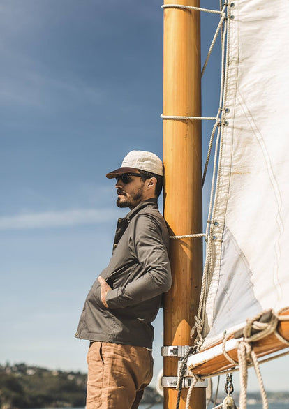 Our guy leaning on a wooden mast wearing a green jacket and brown-peaked cream cap.