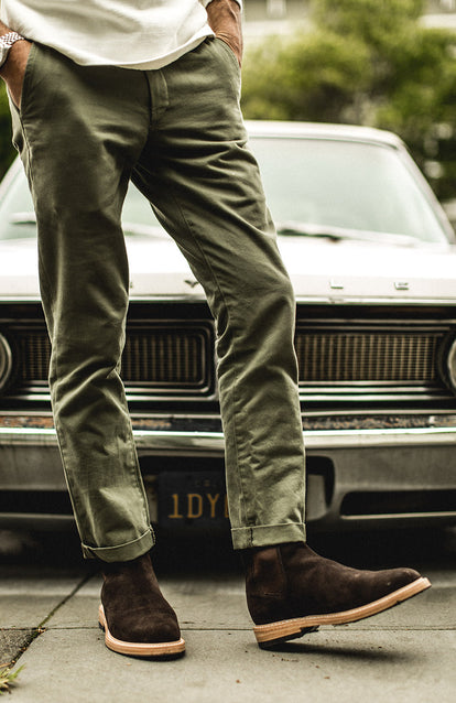 Showing off new suede boots, wearing green chinos, in front of a beat up old car.