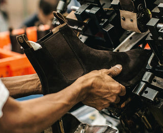 Two hands hold a dark brown suede chelsea boot while a worn metal machine on the right grips its toe.