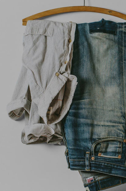 Against a white background, a wooden hanger secures a pair of faded denim jeans and a white and blue striped button down shirt.