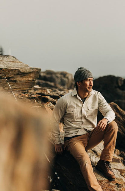 A man in a natural-colored jacket, camel-colored trousers, boots, and a grey beanie sits atop a rock formation and gazes out into the distance.