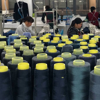 Dozens of spools of dark-colored thread frame a group of four women working at sewing machines in a factory.