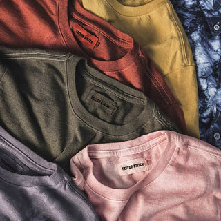 Five T-shirts (each bearing a label with the text Taylor Stitch): one blue, one pink, one olive green, one red, and one yellow, laid out one on top of the next.