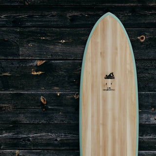 A cedar-colored wooden surfboard with a seafoam-colored outer rim leans against a dark wooden wall.