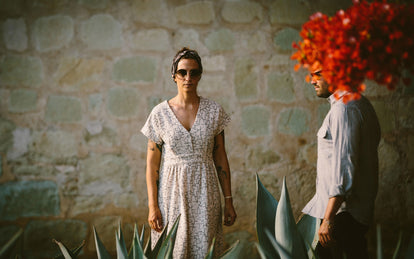 A woman facing the camera, wearing a dress, standing against a stone wall with a man approaching from the right.