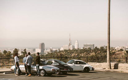 Four or five guys, standing around their parked classic cars, talking, with the city in the far distance.