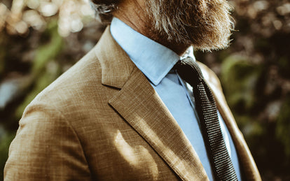 A bearded guy wearing a blue shirt under a beige suit jacket.