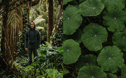Mike Armenta deep in a wet forest, surrounded by greenery, hood up, getting rained on.