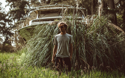 Our guy standing in tall grass, in front of an overgrown trailered cabin cruiser.