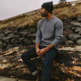 fit model wearing The Adirondack Sweater in Blue Melange sitting on a washed-up log