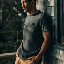 fit model wearingThe Embroidered Heavy Bag Tee in Grey Bear, leaning against house