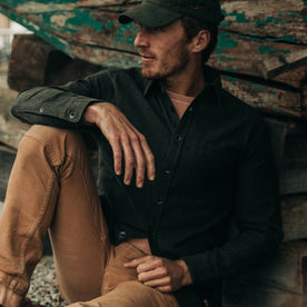 fit model wearing  The Mechanic Shirt in Dark Olive Herringbone, hat on—sitting near boat