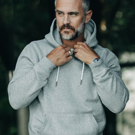 our fit model wearing The Heavy Bag Hoodie in Heather Grey Fleece pulling collar with drawstrings of hood visible