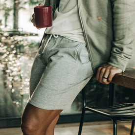 our fit model wearing The Heavy Bag Short in Heather Grey Fleece leaning against the table showing closeup of left side pocket from the front