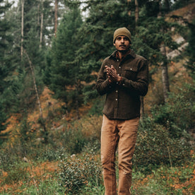 our fit model wearing The Leeward Shirt in Olive Donegal standing in a field with entire shirt visible in front of a forrest