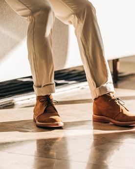 The Unlined Chukka in Butterscotch Weatherproof Suede—pants cuffed, sitting against couch, Footwear by Taylor Stitch